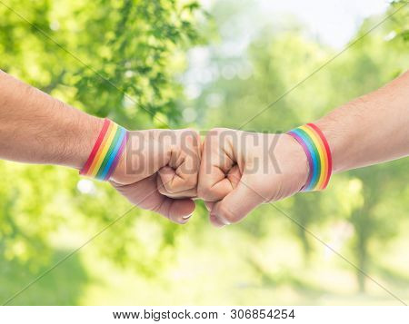 lgbt, same-sex love and homosexual relationships concept - close up of male couple hands with gay pride rainbow awareness wristbands making fist bump gesture over green natural background