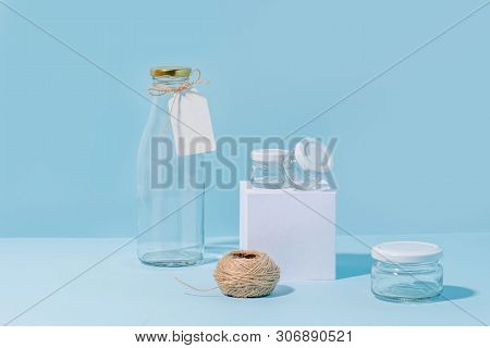 Composition Of Glass Jars On Pedestal, Tagged Empty Milk Bottle And Hank Of Twine Over Blue Backgrou
