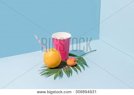 Paper Cup, Fresh Apricot And Orange With Straw Sticking From It Lying Over Palm Branch In The Corner