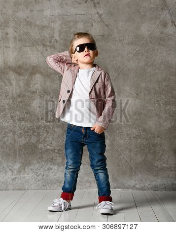 Small Boy Dandy In Light Jeans And A Stylish Jacket, In Sunglasses On The Background Of A Concrete W