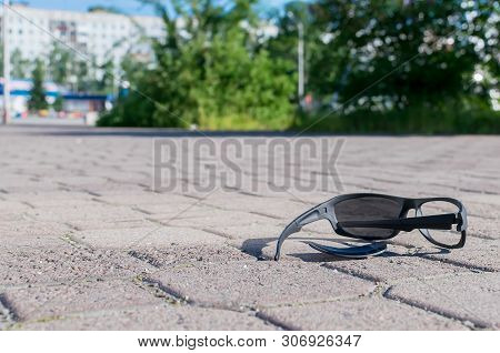 Broken Sun Glasses Lie On A Stone Tiled Walkway In The City Park Against The Background Of Residenti