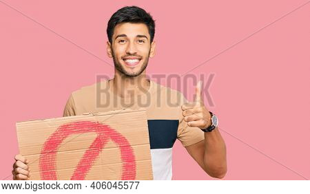 Young handsome man holding prohibited warning banner smiling happy and positive, thumb up doing excellent and approval sign 