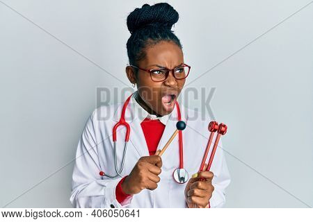 Young african american woman wearing doctor uniform holding tuning fork angry and mad screaming frustrated and furious, shouting with anger. rage and aggressive concept. 
