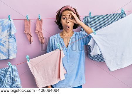 Young brunette woman doing laundry around string hangs doing ok gesture shocked with surprised face, eye looking through fingers. unbelieving expression. 