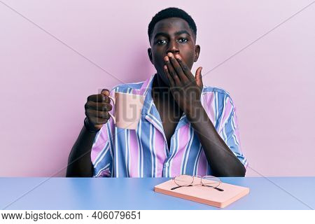 Young african american man sitting on the table reading a book and drinking a coffee covering mouth with hand, shocked and afraid for mistake. surprised expression 