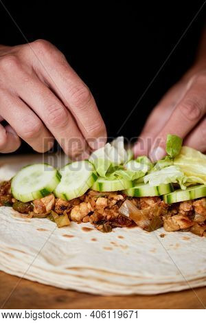 detail of a young caucasian man preparing a durum or a burrito, with chicken meat cooked with different vegetables such as onion or red and green pepper, and fresh lettuce and raw cucumber
