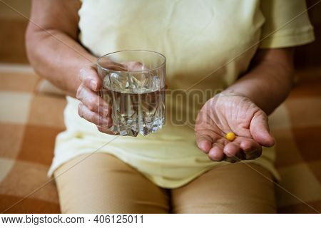 Mature Woman With A Glass Of Water Holds A Tablet In Her Hand While Sitting On The Couch. Treatment 