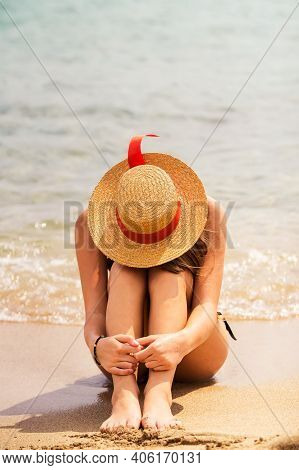 Beautiful Young Tanned Woman In Swim Suit And Straw Hat Sitting At Beach, Put Her Head On Her Legs, 