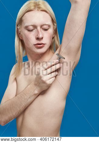 Young Caucasian Man With Long Blond Hair Shaving Armpit With Razor Blade Standing On Blue Background