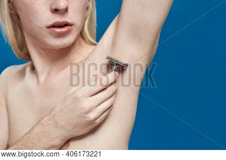 Young Caucasian Man With Long Fair Hair Using Razor For Armpit Shaving While Standing On Blue Backgr