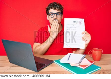 Young caucasian man with curly hair sitting on the table showing failed exam covering mouth with hand, shocked and afraid for mistake. surprised expression 