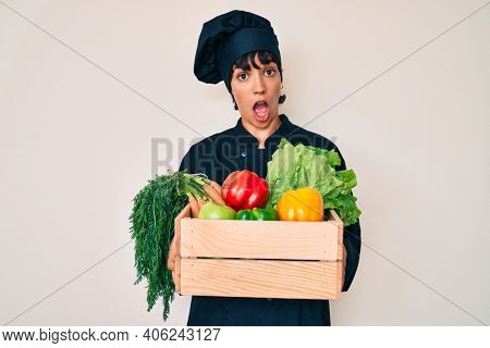 Beautiful brunettte woman chef holding fresh veggetables in shock face, looking skeptical and sarcastic, surprised with open mouth 