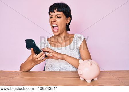 Beautiful brunettte woman caculating money savings angry and mad screaming frustrated and furious, shouting with anger looking up. 