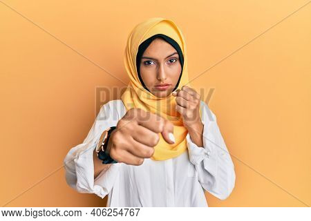 Young brunette arab woman wearing traditional islamic hijab scarf punching fist to fight, aggressive and angry attack, threat and violence 