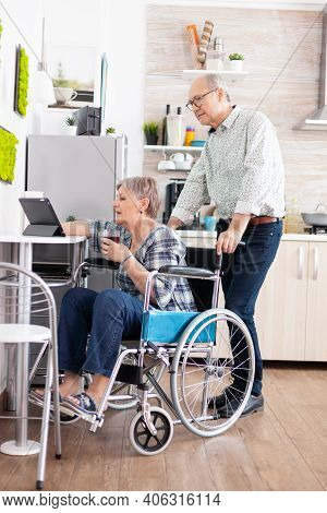 Paralysied Senior Wife In Wheelchair Working At Tablet Computer, Showing To Husband Her Project Sitt
