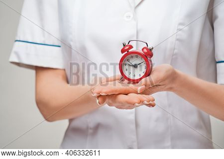 Smiley Nurse Or Doctor Pointing Holding Small Red Alarm Clock Satanding Over Grey Background