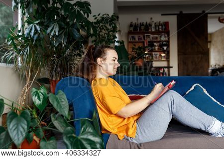 Satisfied And Happy Smiling Girl Sitting On The Sofa In The Room And Writes A Journal Of Your Dreams