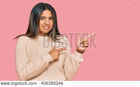 Young latin girl wearing wool winter sweater pointing aside worried and nervous with both hands, concerned and surprised expression 