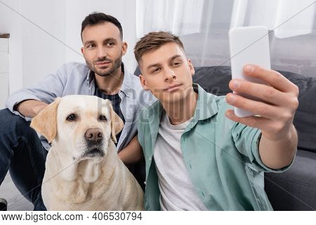 Same Sex Couple Taking Selfie On Smartphone Near Labrador In Living Room.