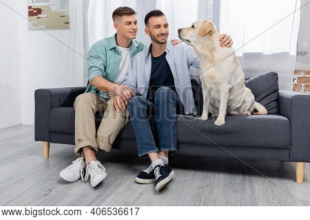 Cheerful Same Sex Couple Smiling And Looking At Labrador In Living Room.