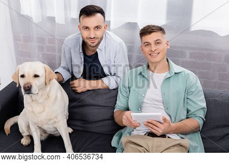 Happy Same Sex Couple Looking At Camera Near Labrador In Living Room.