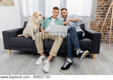 Cheerful Same Sex Couple Watching Movie On Laptop Near Labrador In Living Room.