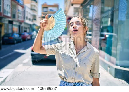 Young beautiful blonde caucasian woman smiling happy outdoors on a sunny day using handfan for hot weather