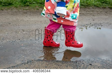A Little Girl In A Bright Jacket And Red Boots Jumps Merrily Through Puddles. Spring Mood.
