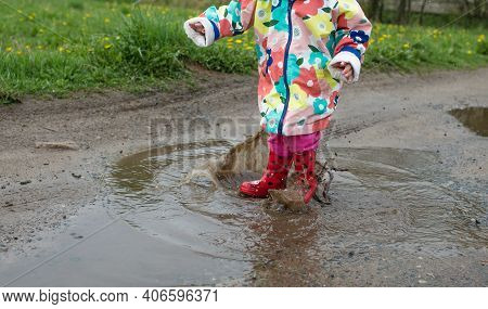 A Little Girl In A Bright Jacket And Red Boots Jumps Merrily Through Puddles. Spring Mood.