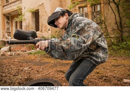 Young Man Playing Paintball. Boy In Camouflage Clothing Aiming A Paintball Gun. Boy In A Protective 