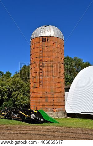 An Old Brown Stone Silo With A Tin Roof And Climbing Ladder Stands Along Side A Barn.