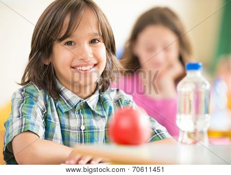 Happy little boy with apple and water bottle sitting in classrom and smiling