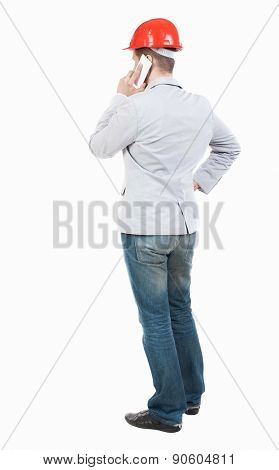 Backview of business man in construction helmet stands and enjoys tablet or using a mobile phone. Standing young guy.  Young businessman talks.