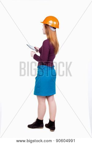 Backview of business woman in construction helmet stands and enjoys tablet or using a mobile phone. Standing young girl. Rear view people collection.  Engineer scorekeeper at work.
