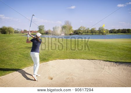 Female golf player hitting ball in sand bunker, with flag on green and lake in background.