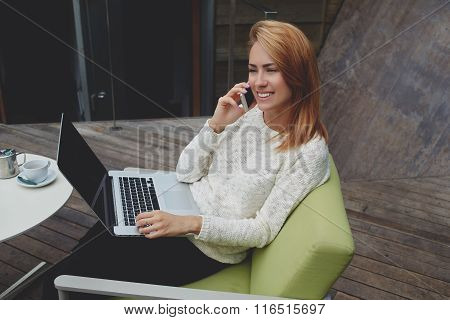 Happy female talking on cell telephone while sitting with laptop computer in cozy cafe