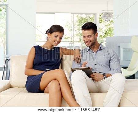 Young caucasian couple at home browsing internet with tablet computer while sitting on sofa. Smiling, pointing at screen, attractive woman.