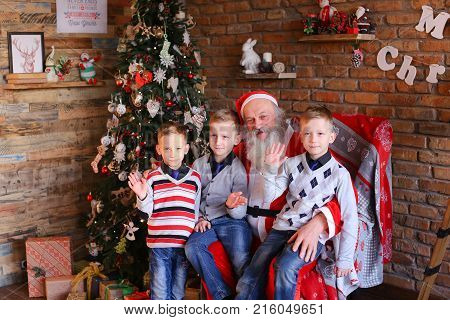 Merry brothers boys sitting on Santa Clauss lap and waving their palms camera rejoice. Claus with gray long beard dressed in red suit white trim pointed cap pompon end. Charming men of European appearance warm sweaters blue jeans socks, sit large soft arm