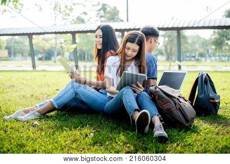 Group of Asian college student using tablet and laptop on grass field at outdoors. Technology and Education learning concept. Future Technology and Modern entertainment concept. Edutainment theme.