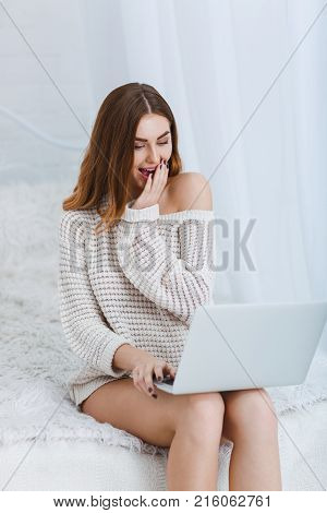 A young, beautiful girl in a white jacket is pleasantly surprised with a laptop kneeling on a bed on a white background