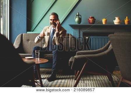Middle-aged Businessman Drinking Coffee And Uses Mobile Phone In The Loby