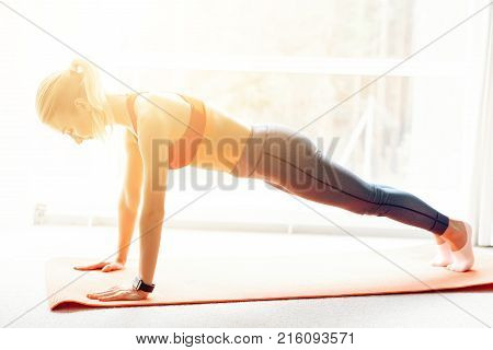 Woman is engaged in yoga in sportswear. She is in a bright room with panoramic windows. She is in kumbhakasana asana, plank pose.