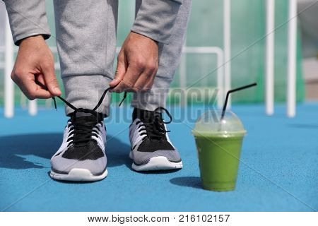 Gym fitness man getting ready tying running shoes with green smoothie detox beverage. Closeup of running trainers with plastic cup of breakfast vegetable juice. Sport active lifestyle.