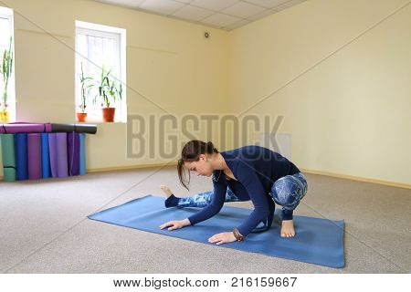 Sportswoman focused on stretch before Pilates class. Girl doing warm up for legs on mat. Female has hair pulled back, concentrated look. Athlete dressed in dark top, yoga legging trains in fitness room. Concept of online training, customizable workouts.