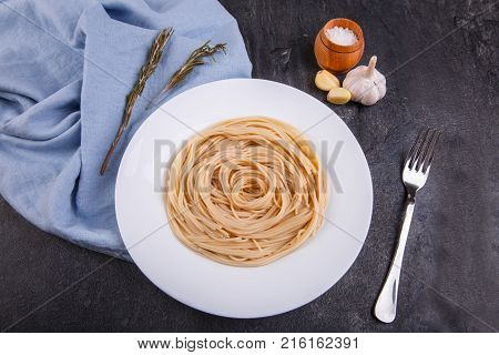 Spaghetti of hard varieties in a white plate, on dark concrete, next to fork, garlic and salt, the plate stands on a blue fabric. View from above.