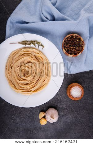 Spaghetti of hard varieties in a white plate, with a sprig of marble, on dark concrete, next to it lies garlic, salt, and picking peppers in peas, a plate stands on a blue fabric. View from above.