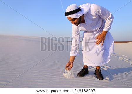 Happy young Emirati Sheikh bends over and takes white sand in hands, rises up and lets sand through fingers of hands, smiling on face, standing in midst of bottomless desert with white sand on hot sunny day. Swarthy handsome Muslim with short dark hair is