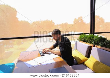 Freelance copywriter typing text fast on laptop keyboard at cafe table and looking tired. Handsome young man dressed in black shirt has dark hair and beard. Concept of typewriting skills and independent contractor for agencies.