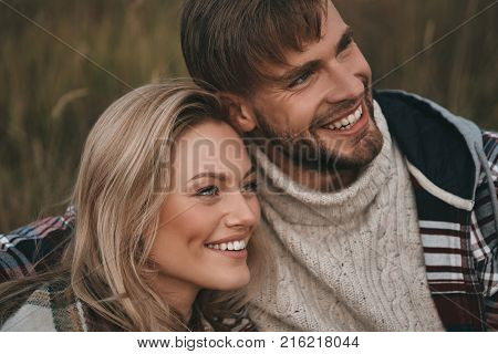 Happy couple. Beautiful young couple embracing and looking away with smile while sitting on the field