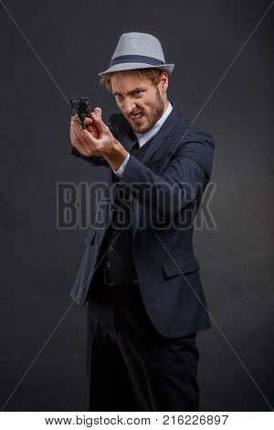 Gangster is stylish and evil in a suit and hat aiming with a gun in his hands on a gray background